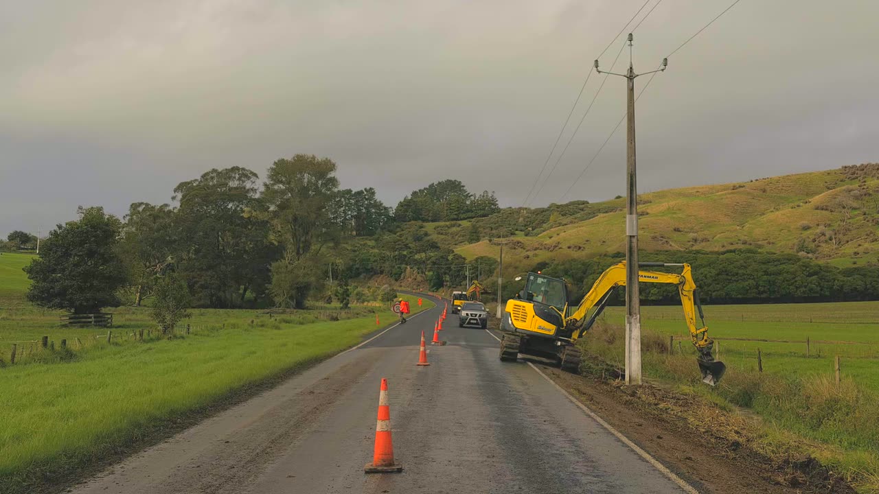 Excavator cutting a drain along the edge of a sealed rural road with cones and a stop go controller