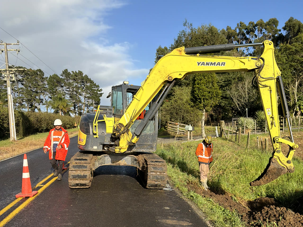 Yanmar tracked excavator on a sealed rural road with its boom over a freshly dug verge drain, two workers in orange hi-vis alongside